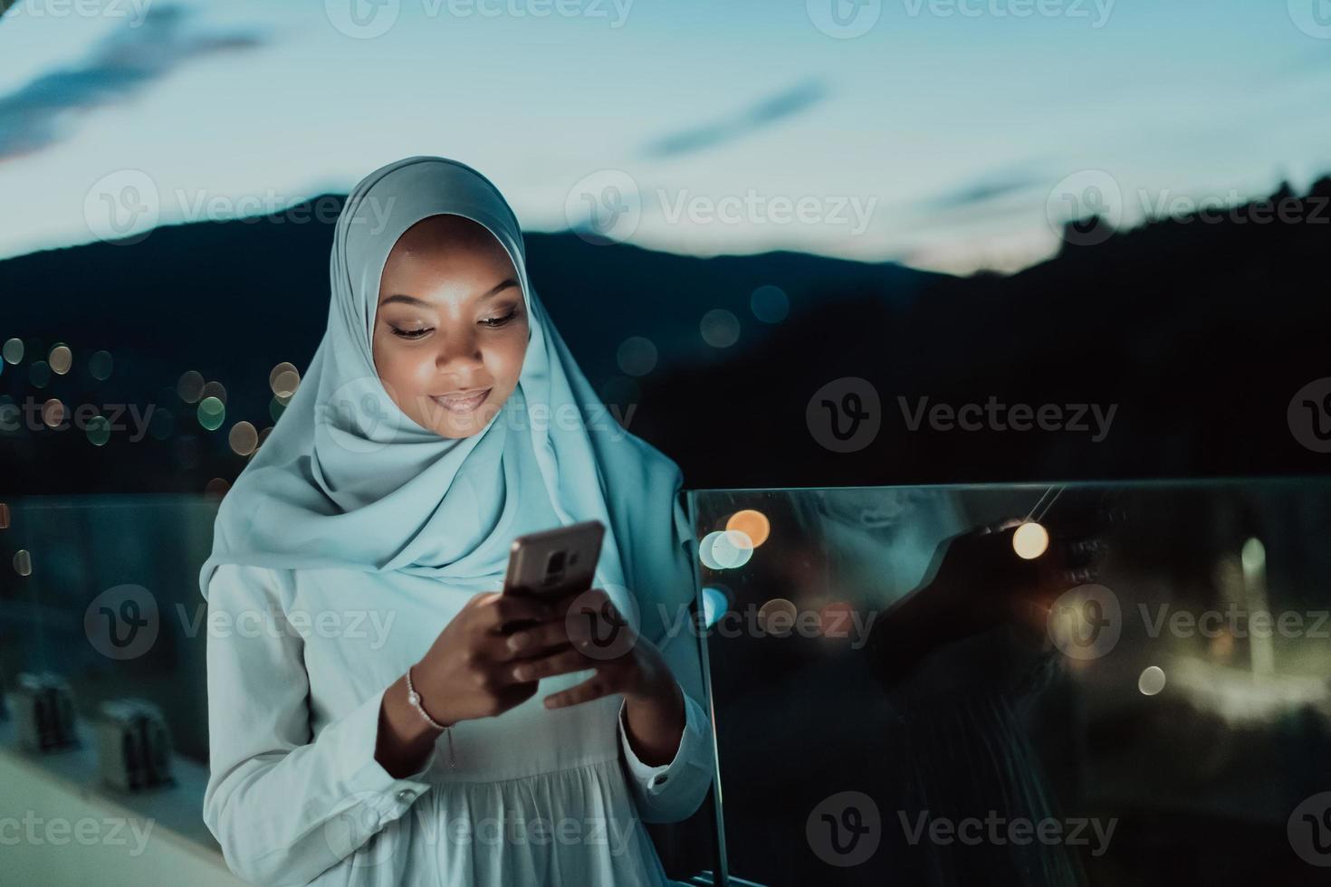 giovane musulmano donna indossare sciarpa velo su urbano città strada a notte sms su un' smartphone con bokeh città leggero nel il sfondo. foto