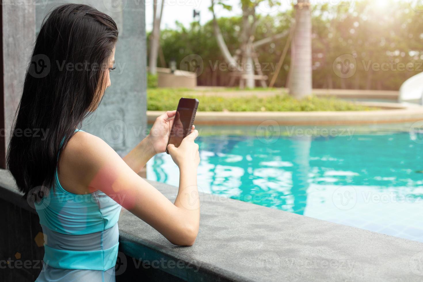 bellissimo carino ragazza indossare blu nuotare completo da uomo bikini, asiatico donna chit Chiacchierare su inteligente Telefono con amico lungo piscina nel ricorrere terme Hotel durante viaggio vacanza vacanze, copia spazio foto