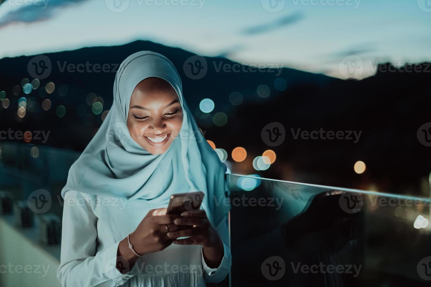 giovane musulmano donna indossare sciarpa velo su urbano città strada a notte sms su un' smartphone con bokeh città leggero nel il sfondo. foto