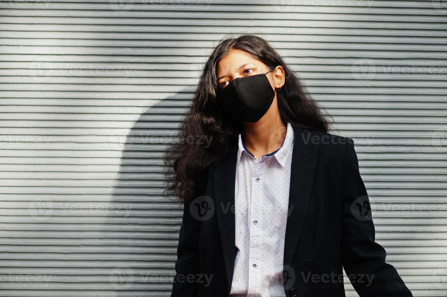 bellissima indiano donna indossare formale e nero viso maschera, in posa contro parete durante covid pandemismo. foto