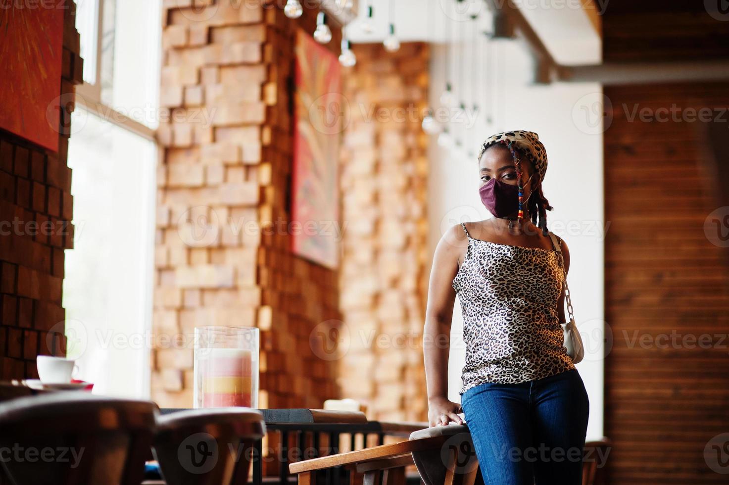 elegante africano americano donna con dreadlocks afro capelli, indossare viso proteggere maschera e copricapo leopardo sciarpa a ristorante. nuovo normale vita dopo coronavirus epidemico. foto