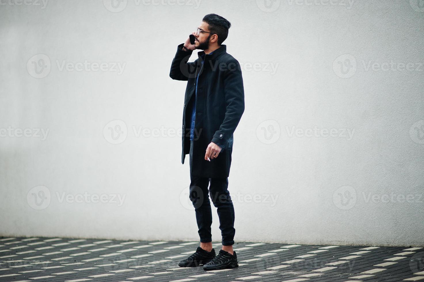 mezzo orientale imprenditore indossare nero cappotto e blu camicia, occhiali in piedi contro bianca parete, fumo sigaretta e parlare di Telefono. foto