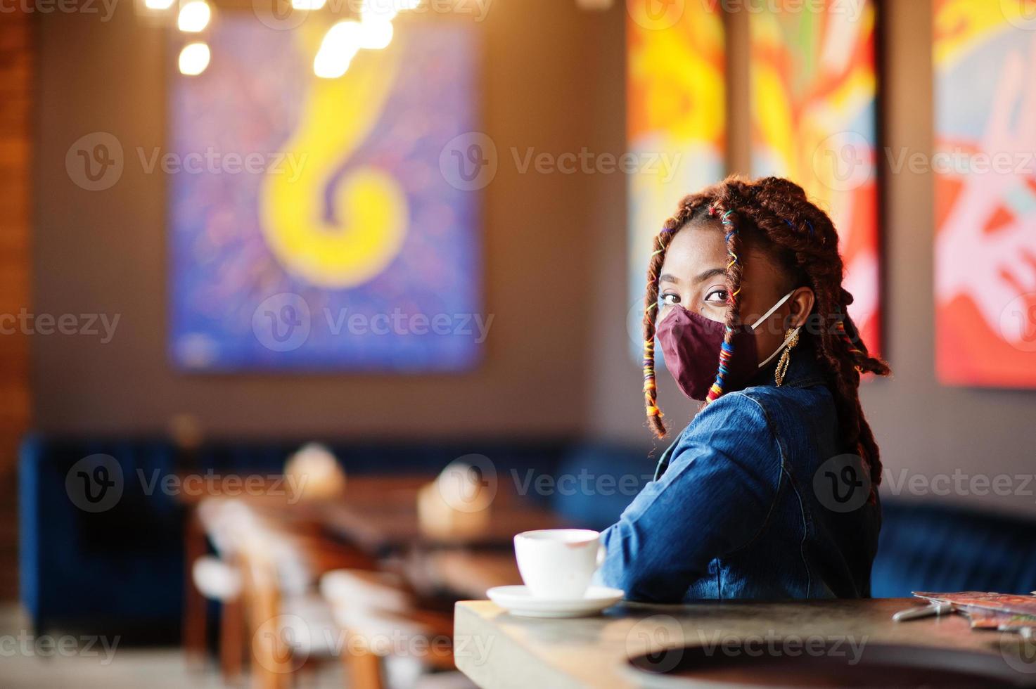 elegante africano americano donna con dreadlocks afro capelli, indossare jeans giacca e viso proteggere maschera a ristorante. nuovo normale vita dopo coronavirus epidemico. foto