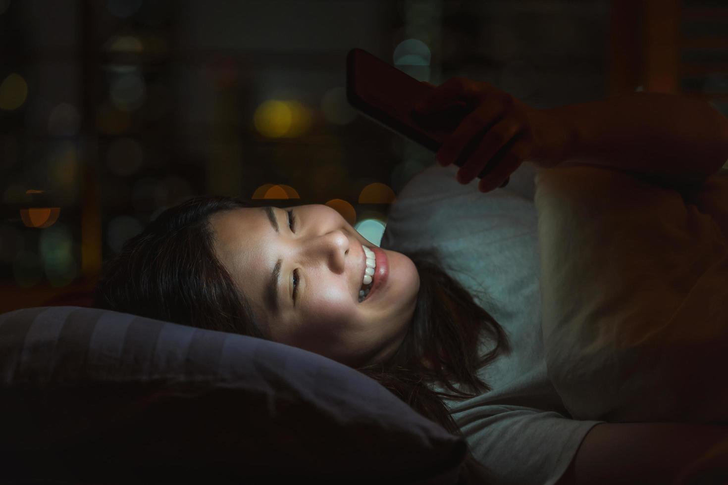 giovane donna asiatica che per mezzo del telefono mobile astuto per affrontare una persona amata prima di andare a letto foto
