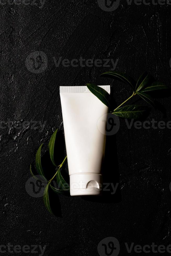 bianca cosmetico tubo con mano crema su un' buio sfondo con verde le foglie. cosmetici concetto con erbaceo ingredienti foto