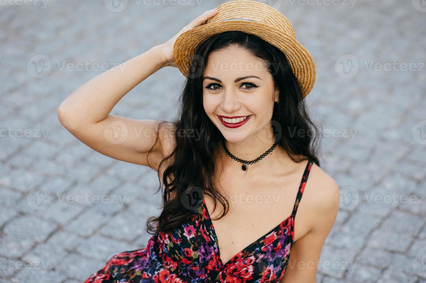 bella donna bruna alla moda con il trucco che indossa un cappello di paglia sulla testa e un vestito estivo che sorride piacevolmente nella fotocamera mentre posa all'aperto. felice bella giovane donna che modella sulla strada foto