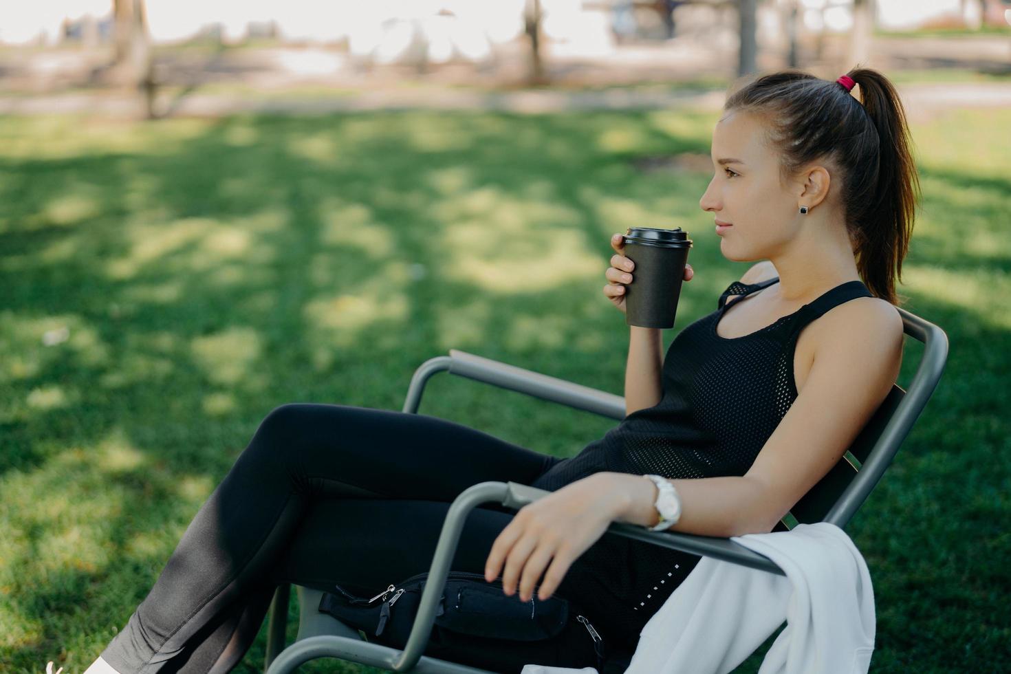 la giovane donna sportiva premurosa con la coda di cavallo ha una pausa caffè dopo l'allenamento pone in una sedia comoda con una bevanda calda nel parco contro l'erba verde gode di una giornata di sole concentrata a distanza foto