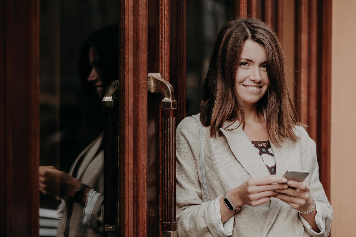 donna allegra e allegra con capelli lisci scuri, usa lo smartphone, vestita con abiti eleganti, guarda positivamente la fotocamera, ama la comunicazione online, controlla il percorso, invia messaggi, legge le notifiche foto