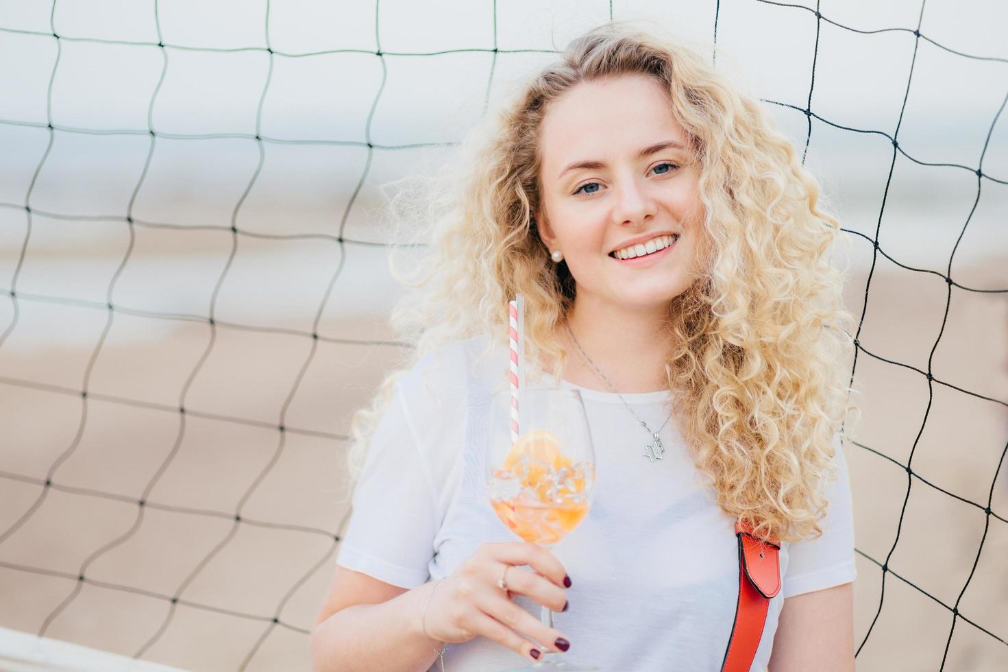 giovane modella ottimista con capelli ricci chiari, vestita con una maglietta bianca casual, beve un cocktail fresco, si gode un buon riposo in spiaggia, si trova vicino alla rete da tennis. concetto di persone, bellezza e riposo foto