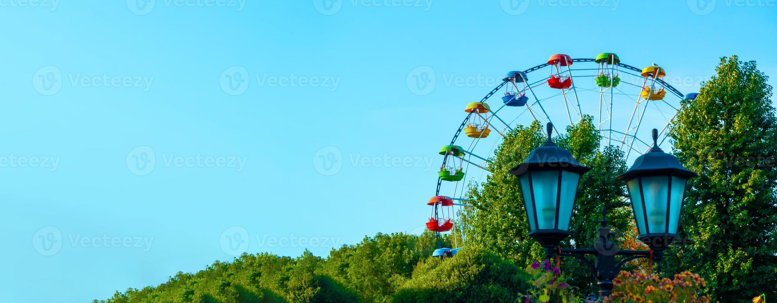 paesaggio di un parco divertimenti con lanterna decorata con fiori sullo sfondo la parte superiore di una ruota panoramica che mostra sopra le cime degli alberi contro un cielo blu. foto