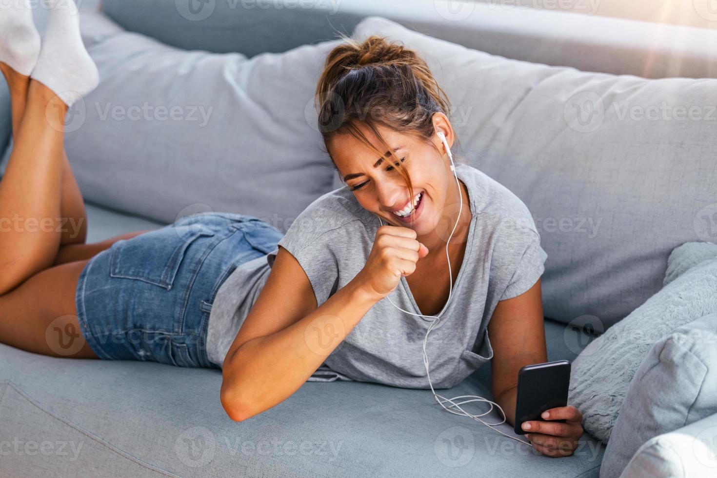 rilassare nel il ritmo di mio musica. attraente giovane donna lcon auricolari e smartphone seduta su divano a casa. bellissimo ragazza nel denim e cuffie è ascoltando per musica utilizzando un' inteligente Telefono foto