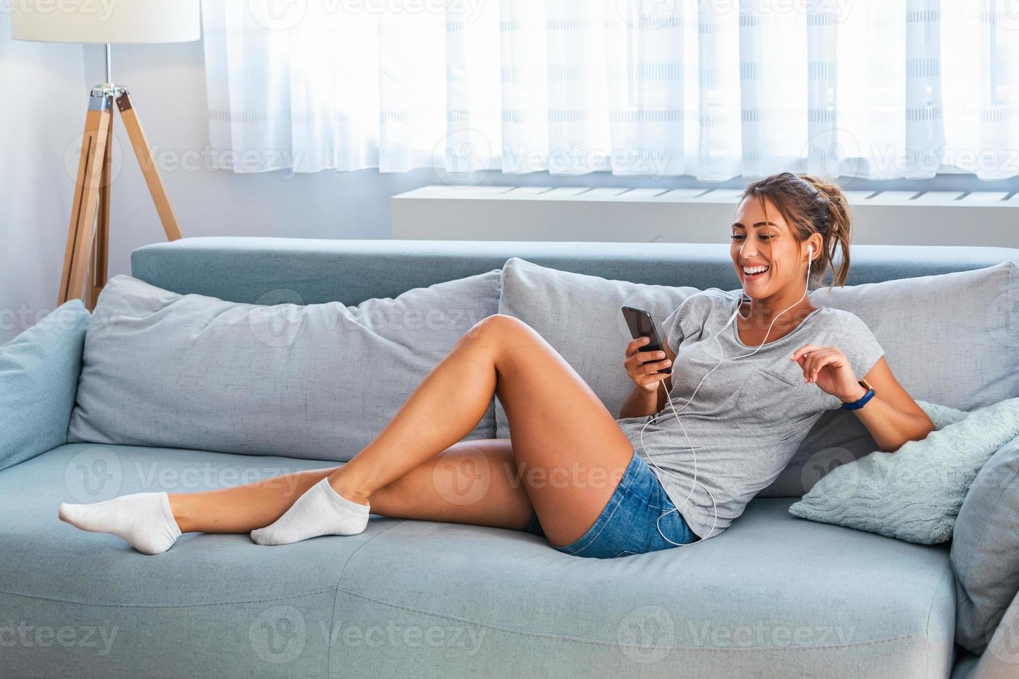 rilassare nel il ritmo di mio musica. attraente giovane donna lcon auricolari e smartphone seduta su divano a casa. bellissimo ragazza nel denim e cuffie è ascoltando per musica utilizzando un' inteligente Telefono foto