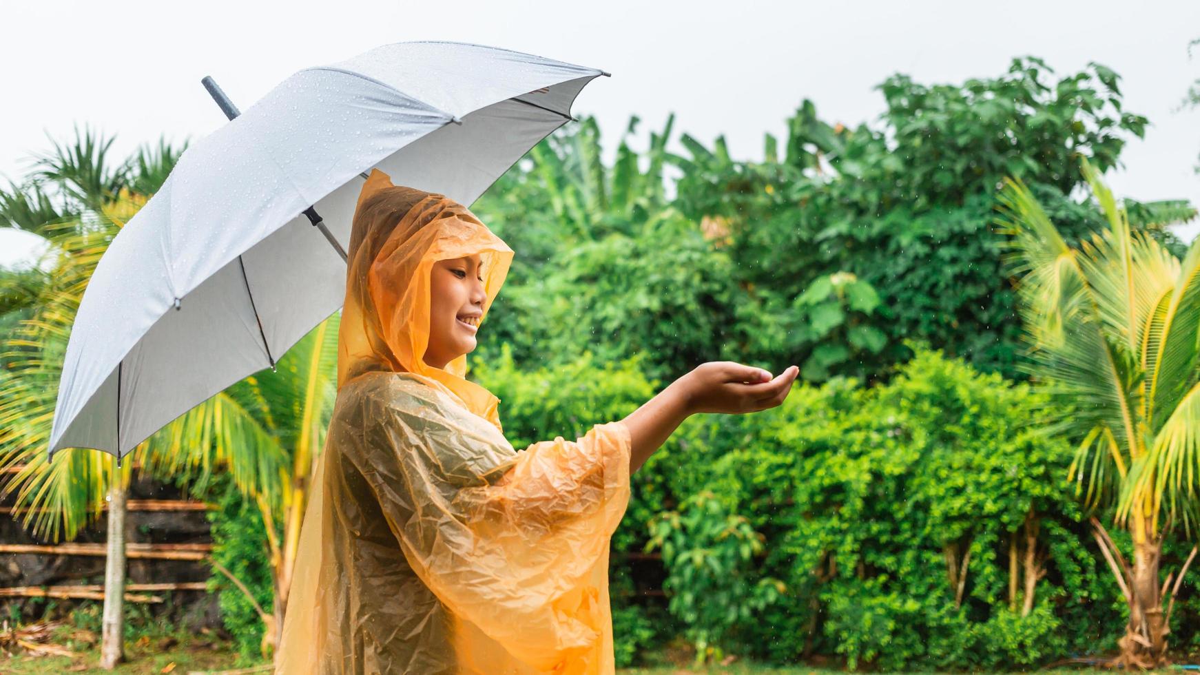 asiatico ragazzo indossare arancia impermeabile Tenere ombrello contento e avendo divertimento nel il pioggia su un' piovoso giorno foto