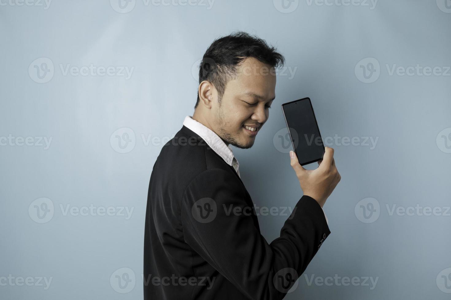 un' ritratto di un' contento asiatico uomo d'affari è sorridente e Tenere il suo smartphone mostrando copia spazio su è schermo indossare nero completo da uomo isolato di un' blu sfondo foto