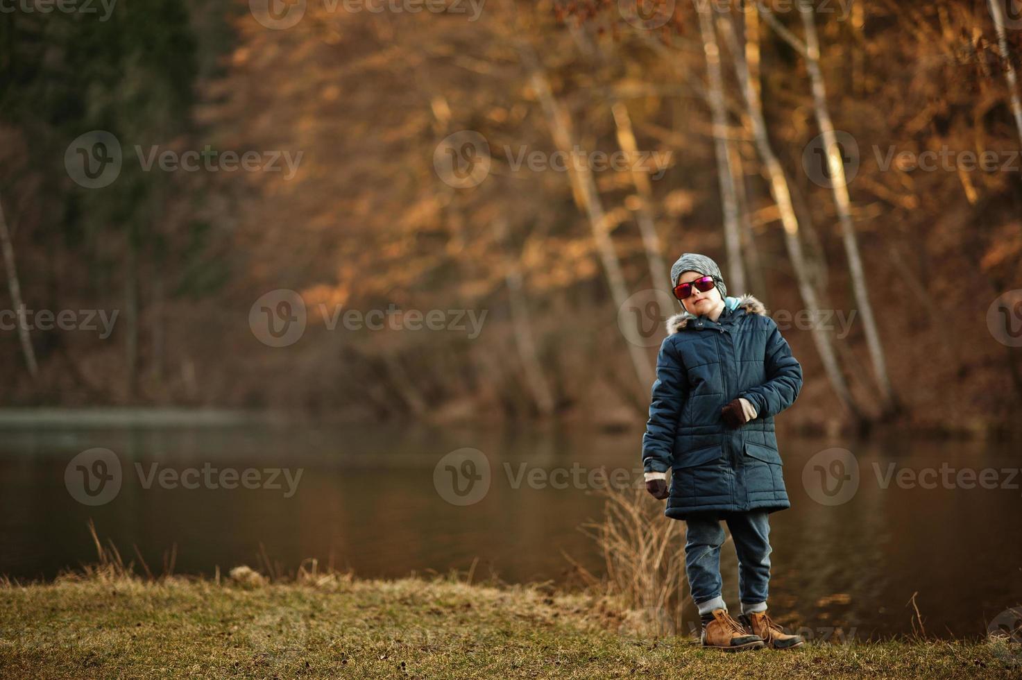 ragazzo nel giacca e occhiali da sole In piedi nel soleggiato primavera parco contro fiume. foto