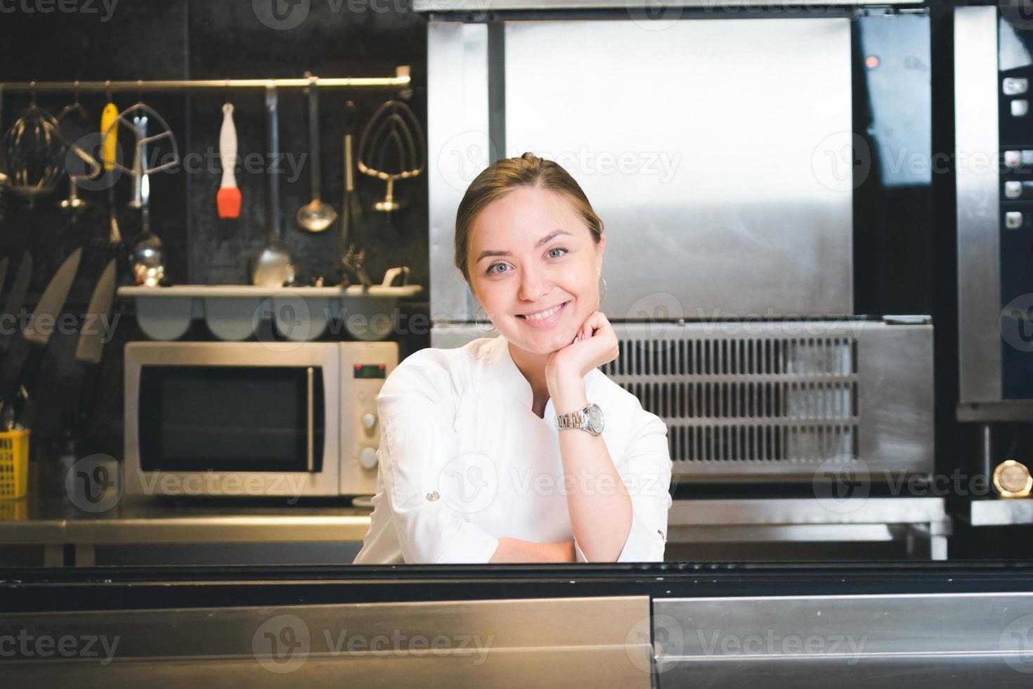 ritratto di fiducioso e sorridente giovane donna capocuoco vestito nel bianca uniforme, professionale cucina siamo su sfondo foto