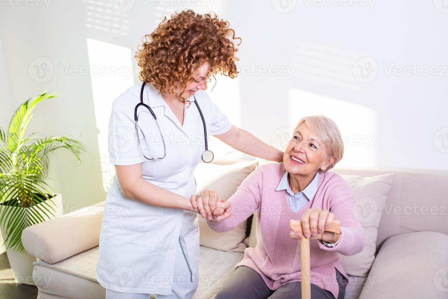 femmina badante porzione anziano donna ottenere su a partire dal divano nel vivente camera. sorridente infermiera assistere anziano donna per ottenere su. cura infermiera supporto paziente mentre ottenere su a partire dal divano foto