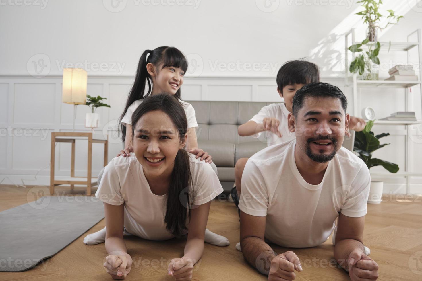 felice benessere famiglia tailandese asiatica, i bambini giocano e prendono in giro i loro genitori mentre l'allenamento fitness yoga e l'esercizio fisico insieme nel soggiorno bianco, lo stile di vita domestico, l'attività del fine settimana. foto