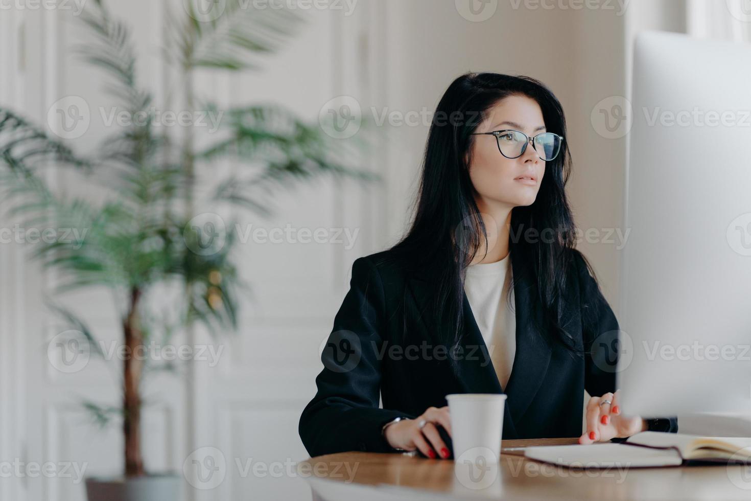 l'imprenditrice concentrata indossa un elegante costume nero, lavora al computer, si siede a tavola in un luogo di lavoro accogliente indossa occhiali trasparenti naviga in internet fa progetti di business pensa alla strategia foto