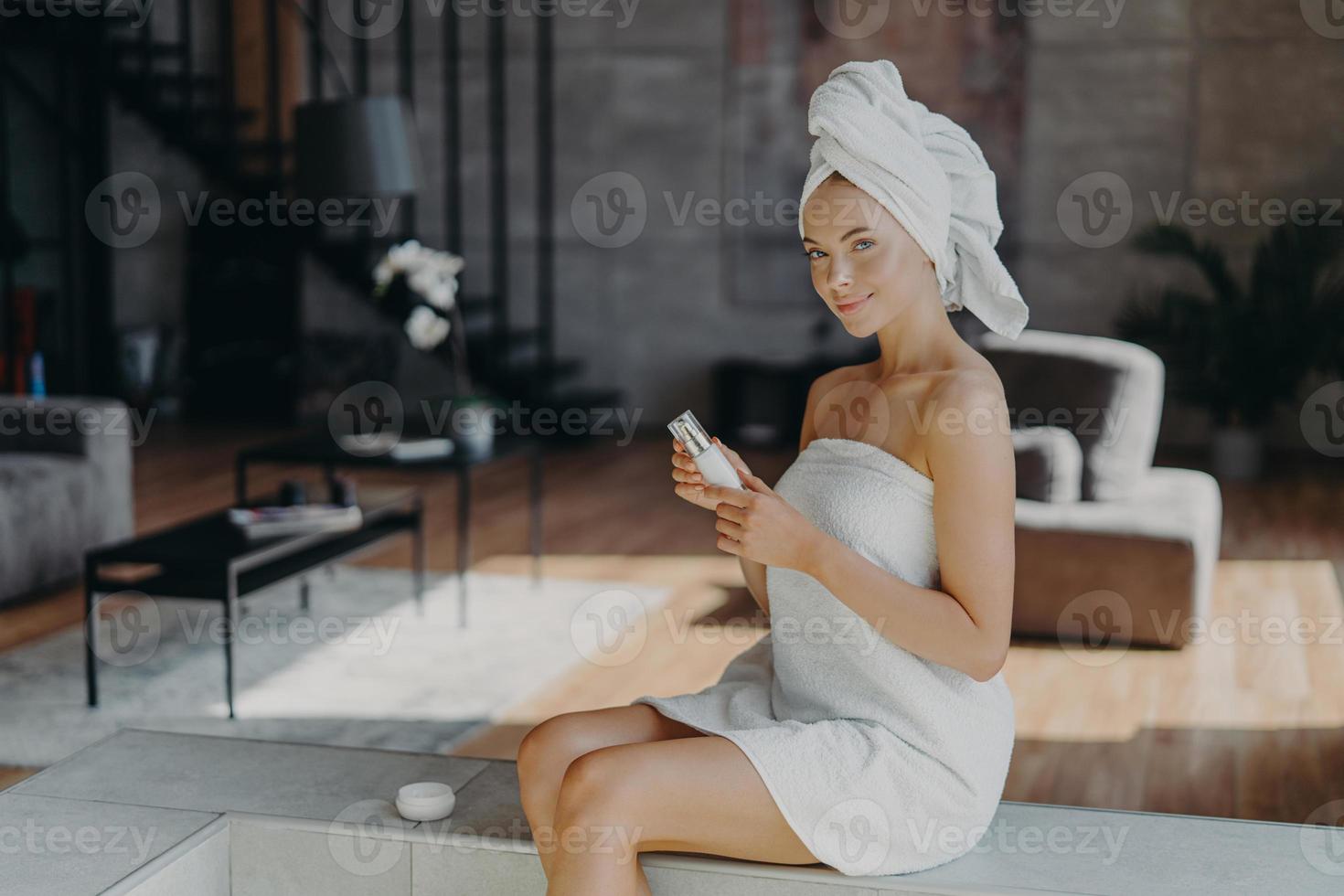 l'inquadratura orizzontale di una bella donna attraente si siede sullo sfondo della stanza accogliente applica la lozione per il corpo dopo aver fatto il bagno usa un prodotto cosmetico per una pelle luminosa avvolta in un asciugamano si sottopone a trattamenti di bellezza foto