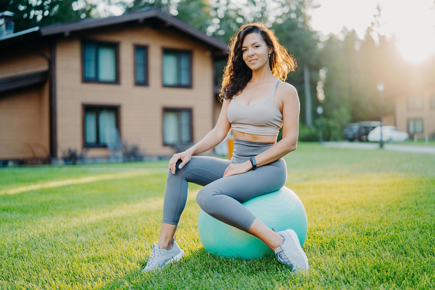 concetto di fitness, sport e allenamento. soddisfatta donna sportiva in buona forma fisica si prende una pausa dopo l'esercizio si siede sulla palla fitness all'aperto gode della natura e dell'aria fresca, fa esercizi di equilibrio pilates foto