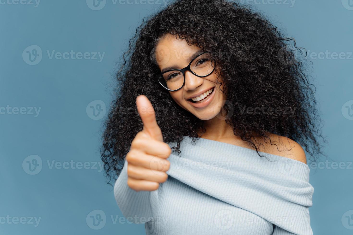 la sua eccellente donna afroamericana sorridente e felice mostra come un gesto, tiene il pollice alzato, dà approvazione, applaude oltre il muro blu, vestita con abiti casual, esprime approvazione per un prodotto straordinario foto