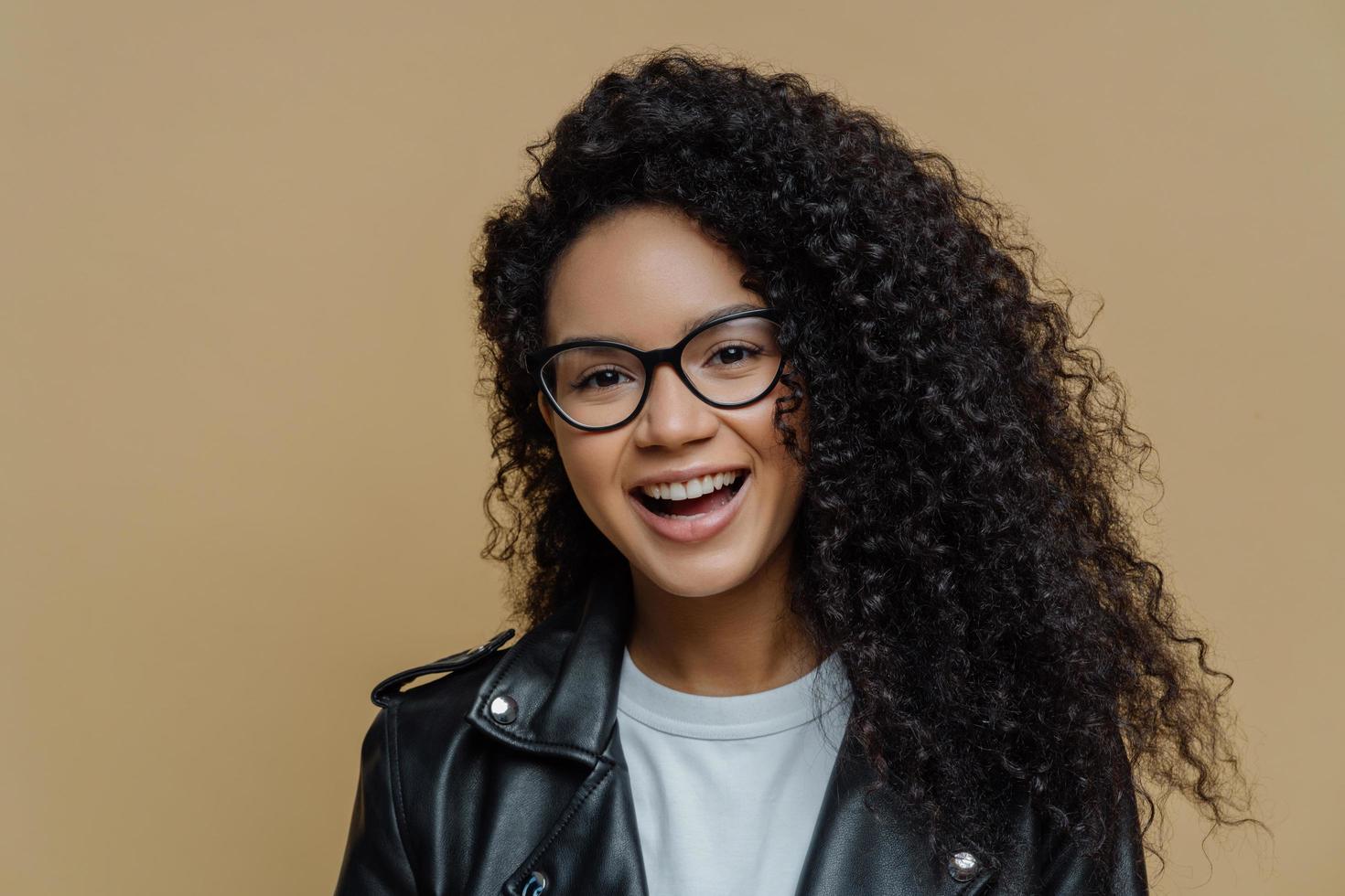 colpo alla testa di una donna afroamericana allegra con capelli ricci scuri, ha un'espressione facciale felice, una conversazione gioiosa, indossa occhiali trasparenti e giacca di pelle nera, isolata su sfondo beige foto