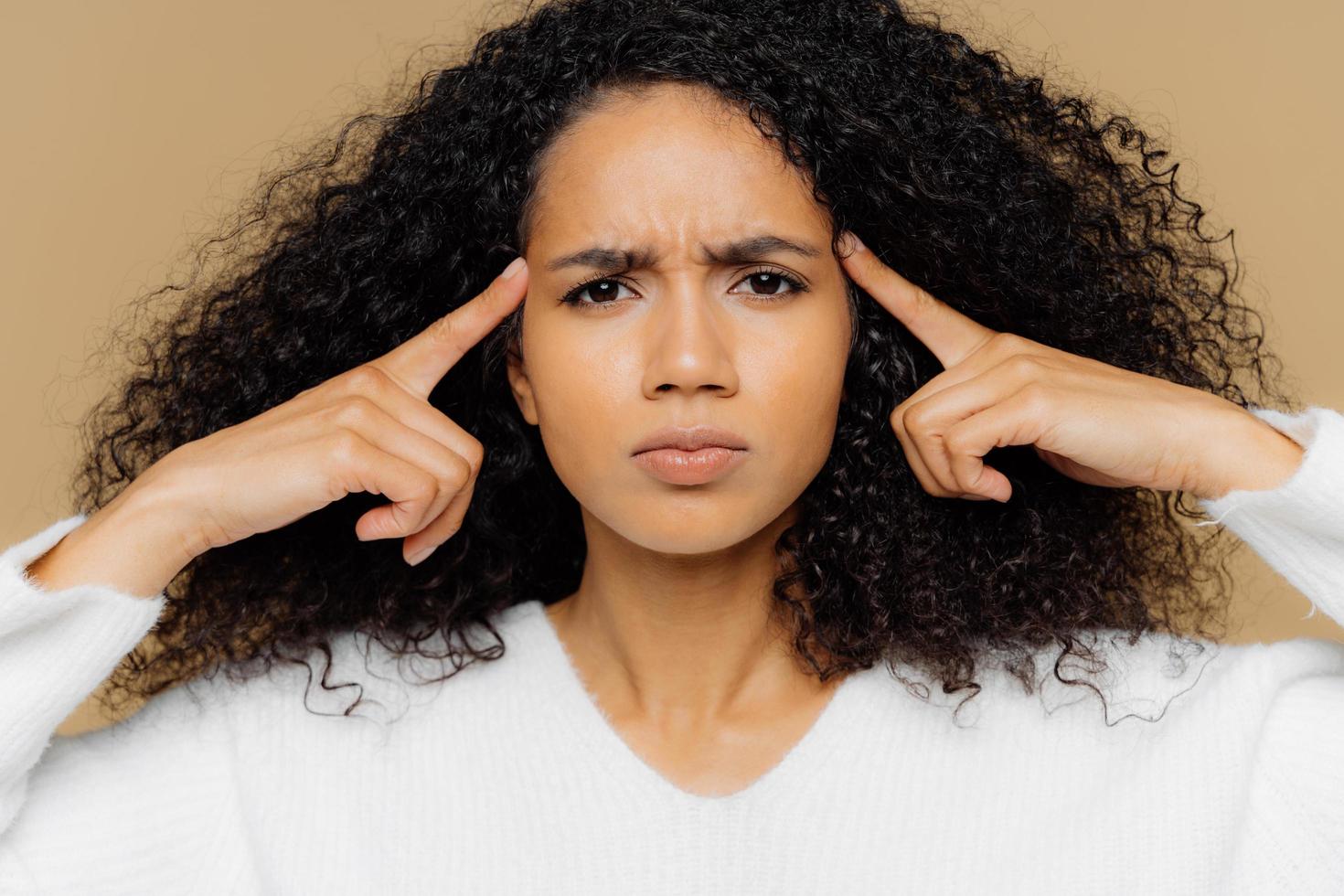 colpo alla testa di una donna afroamericana stressante tiene gli indici sulle tempie, soffre di mal di testa aggrotta le sopracciglia, ha un'espressione facciale insoddisfatta, vestita in modo casual, modella al chiuso. persone ed emicrania foto