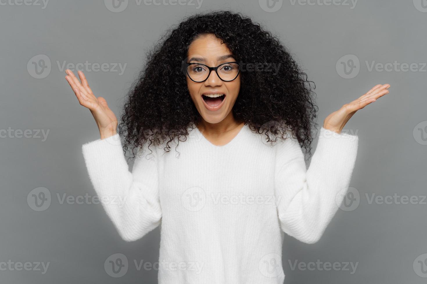 la donna afroamericana soddisfatta alza le mani nel gesto eureka, tiene i palmi delle mani sollevati, la bocca spalancata, indossa occhiali da vista e un maglione bianco, isolato su sfondo grigio. wow, è fantastico. foto