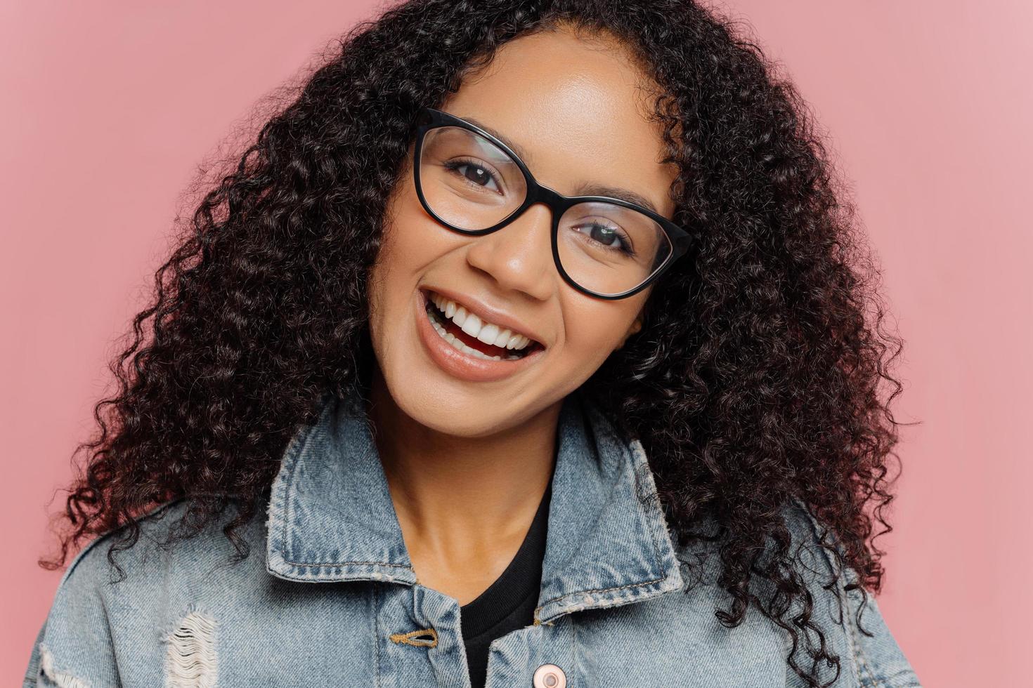 primo piano ritratto di donna sorridente felice con acconciatura afro riccia scura, testa inclinata, indossa occhiali ottici e giacca di jeans, isolata su sfondo roseo. persone, etnia, emozioni e sentimenti foto