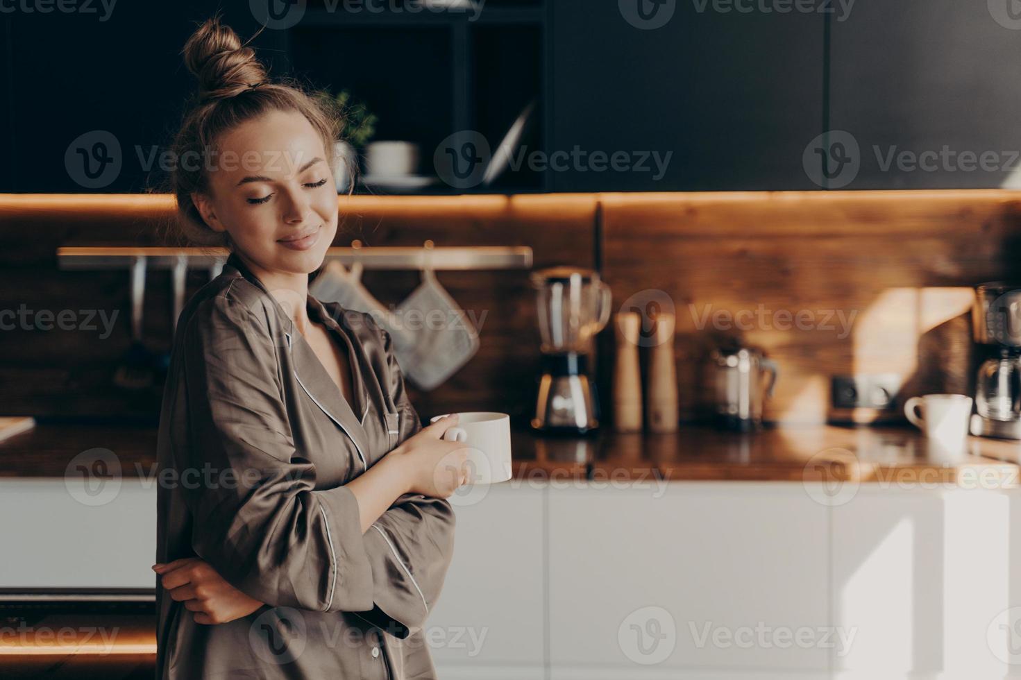 bella giovane donna felice in piedi in un accogliente pigiama di seta in cucina con una tazza di caffè foto