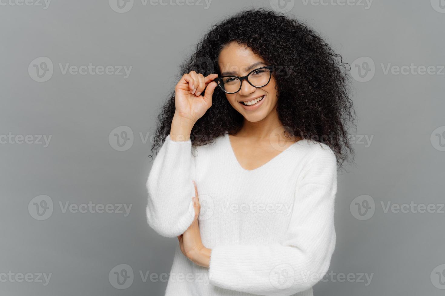 ritratto di contento donna con Riccio capelli, conserva mani su telaio di occhiali, ha dolce Sorridi, indossa bianca Maglione, isolato al di sopra di grigio sfondo. copia spazio per il tuo annuncio. Contentissimo alunno interno foto