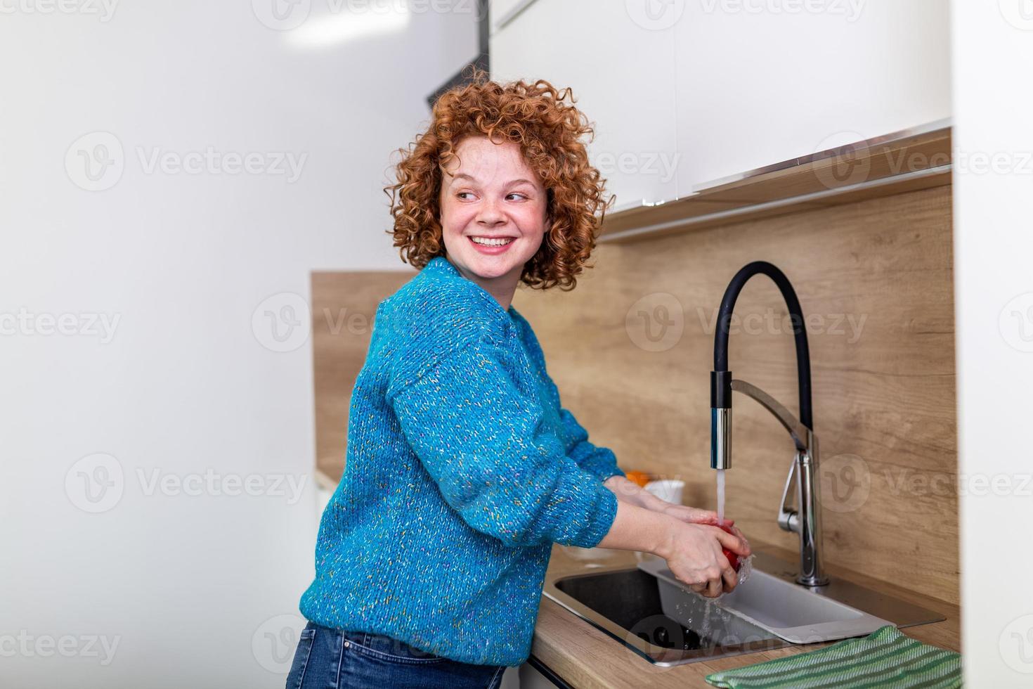 carino ragazza con Riccio rosso capelli lavaggio Mela nel cucina lavello. giovane donna pulizia sua frutta con acqua sotto il rubinetto. quotidiano presa di vitamine con frutta, dieta e salutare mangiare foto