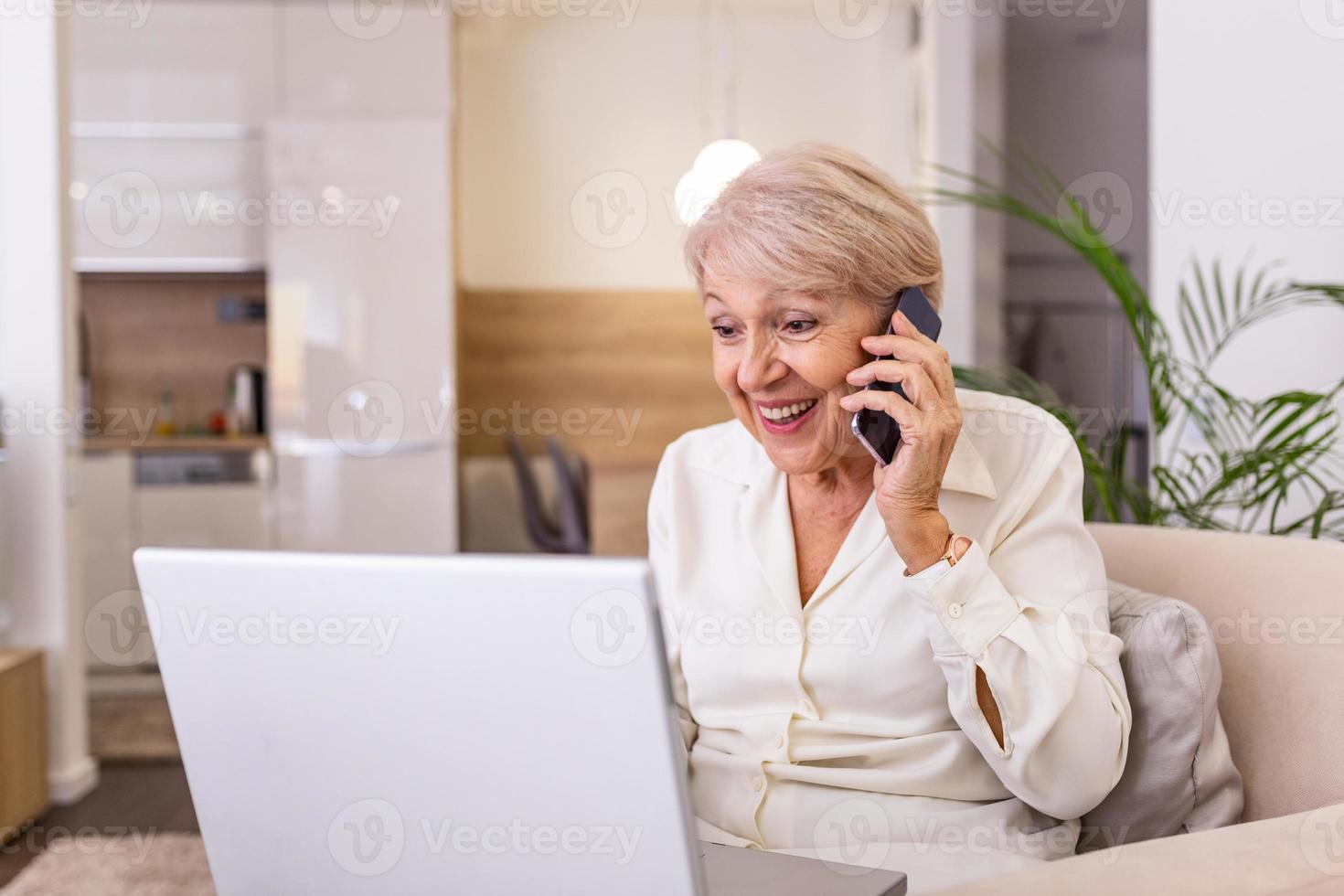 anziano donna Lavorando su il computer portatile computer, sorridente, parlando su il Telefono. anziano donna utilizzando il computer portatile. anziano donna seduta a casa, utilizzando il computer portatile computer e parlando su sua mobile Telefono, sorridente. foto