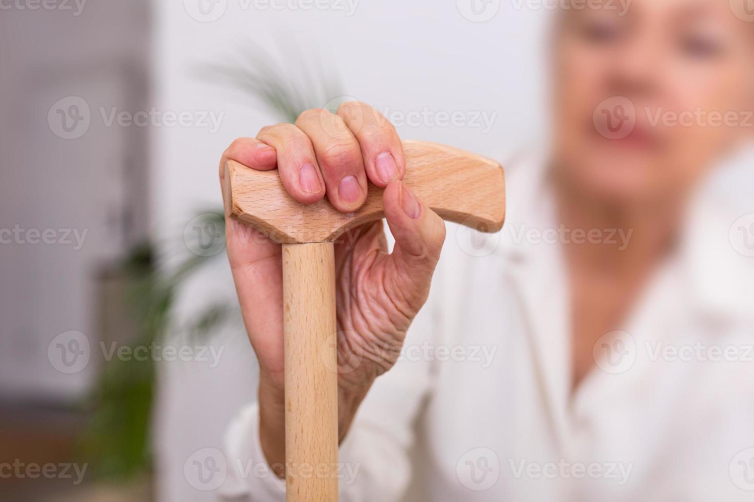 mani di un vecchio donna con un' canna, Sambuco signora seduta su il divano con di legno a piedi bastone. ritagliata tiro di un' anziano donna Tenere un' canna nel un' la pensione casa foto