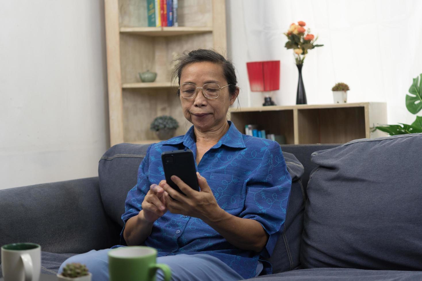 anziano donna asiatico Tenere cellula Telefono su divano nel casa. attività commerciale comunicazione e Internet sociale networking tecnologia. foto