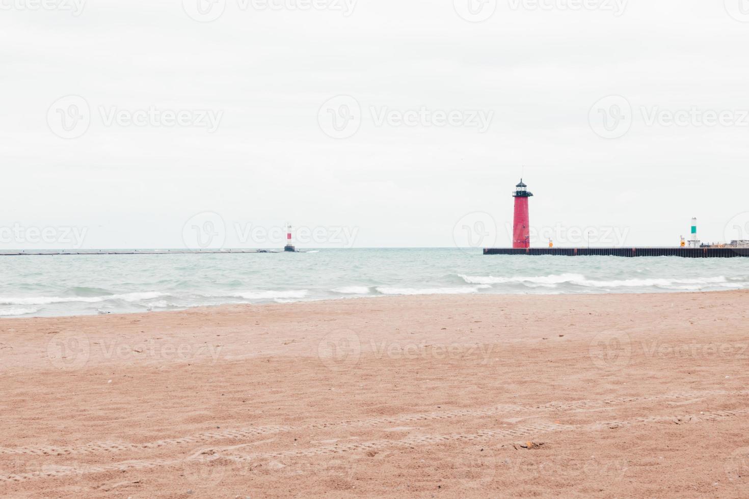 grigio nuvoloso cielo di lago Michigan. onde rotolamento in il vuoto spiaggia. rosso faro visto a il fine di il molo per avvisare diportisti. boe visto nel il distanza. impronte nel il sabbia. foto
