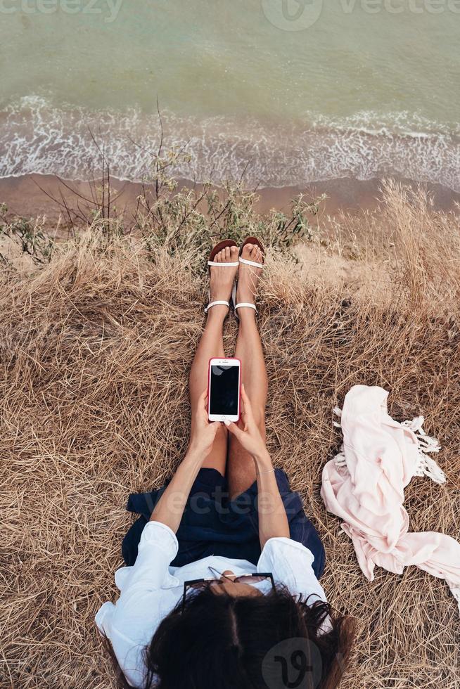 bellissimo giovane ragazza seduta e Tenere un' smartphone nel sua mani, superiore Visualizza foto