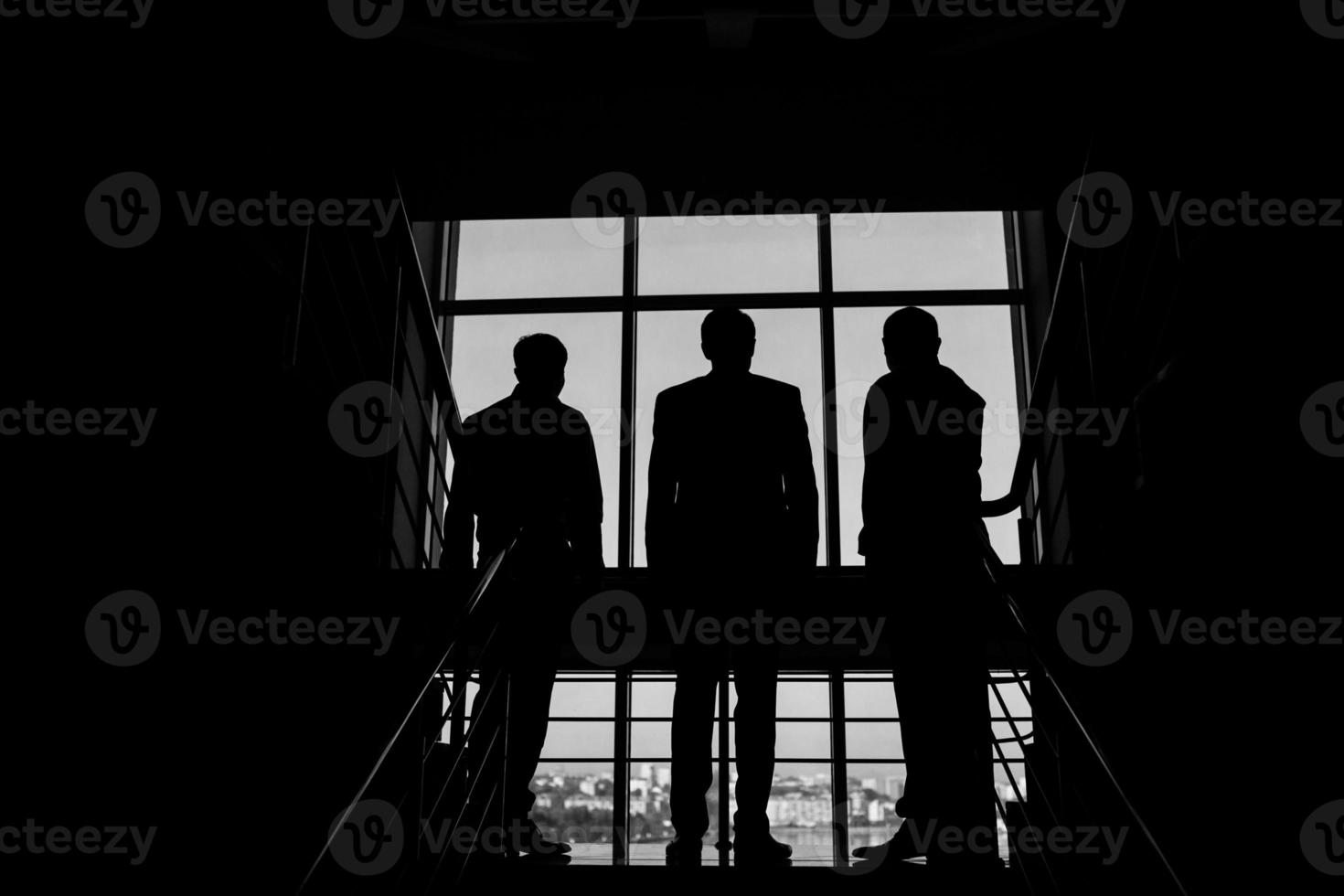 tre uomini d'affari in piedi a il finestra con un' città Visualizza foto