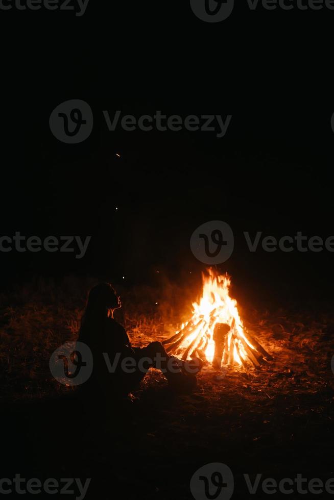 donna seduta e ottenere caldo vicino il falò nel il notte foresta. foto