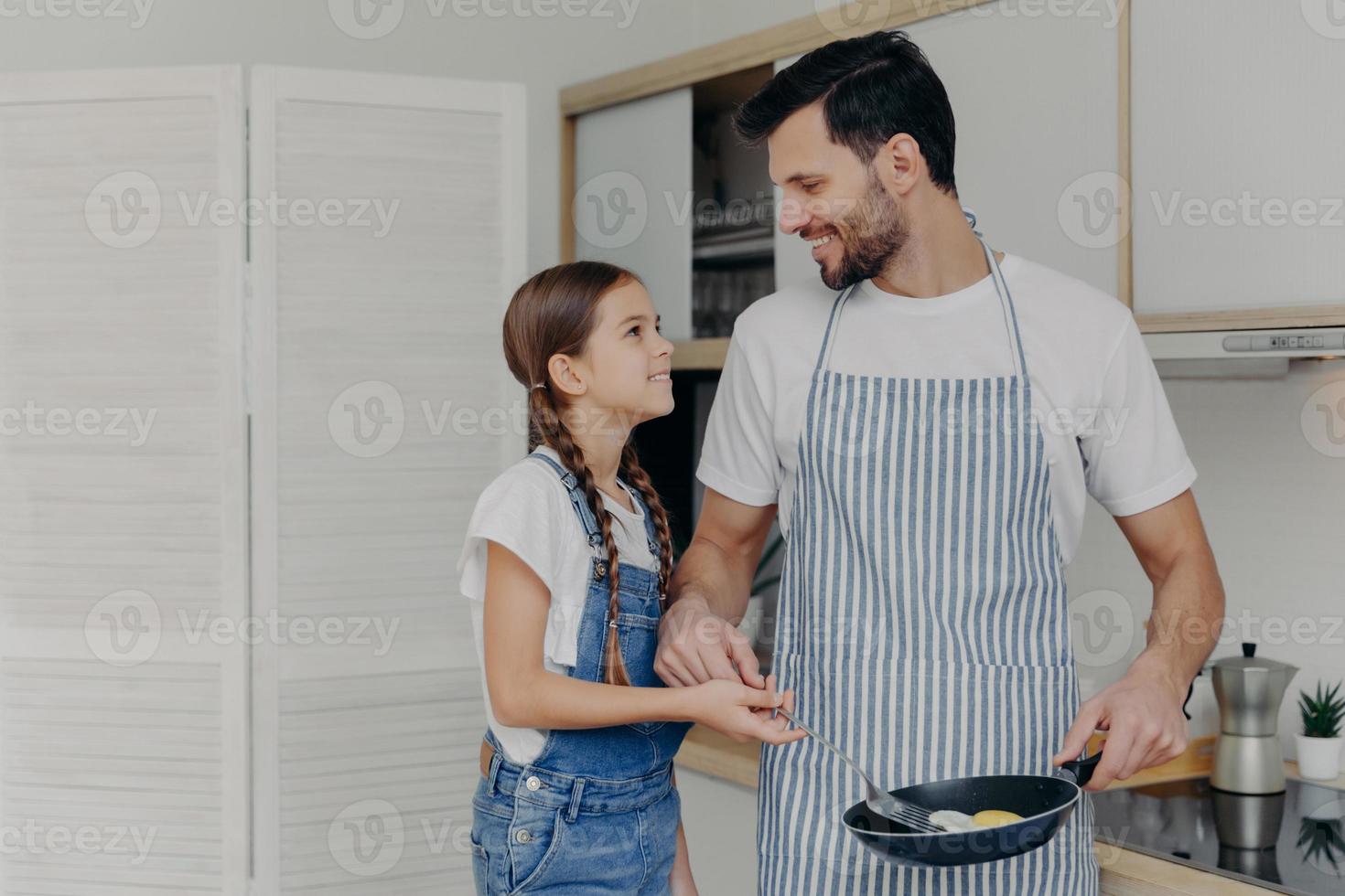 padre affettuoso in apton frigge le uova, insegna alla figlia piccola come cucinare, posa insieme in cucina, prepara una deliziosa colazione, guardati con un sorriso. figli, paternità, vita domestica foto