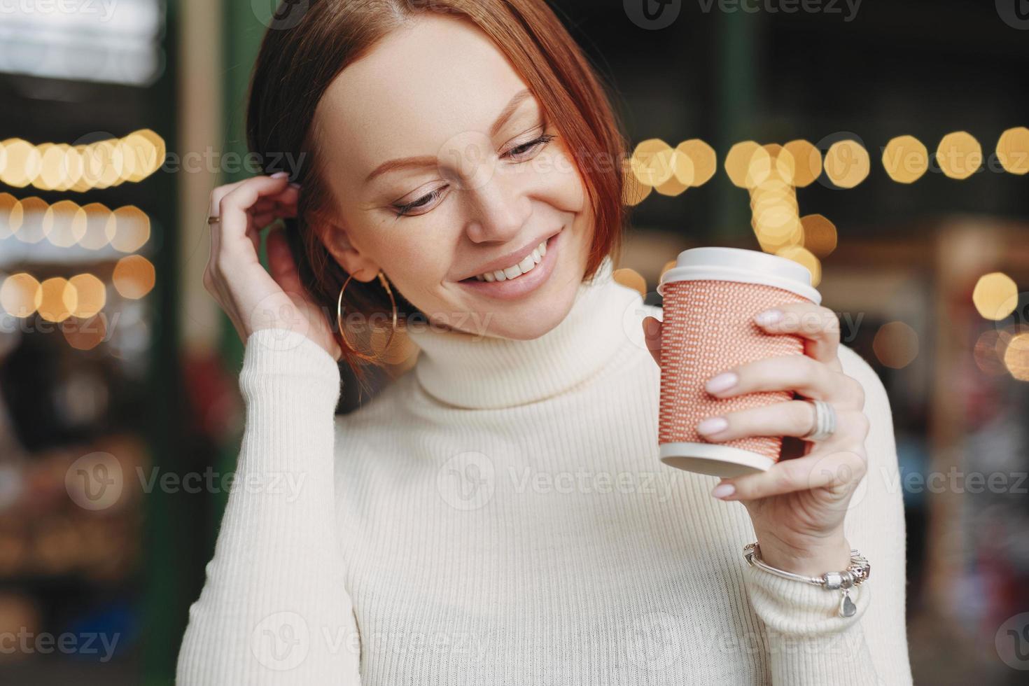 foto ritagliata di una donna dall'aspetto piacevole posa in un caffè all'aperto, tiene una tazza di caffè di carta, vestita con un abito bianco, sorride felicemente, parla con un collega o un amico durante la pausa. femmina con bevanda