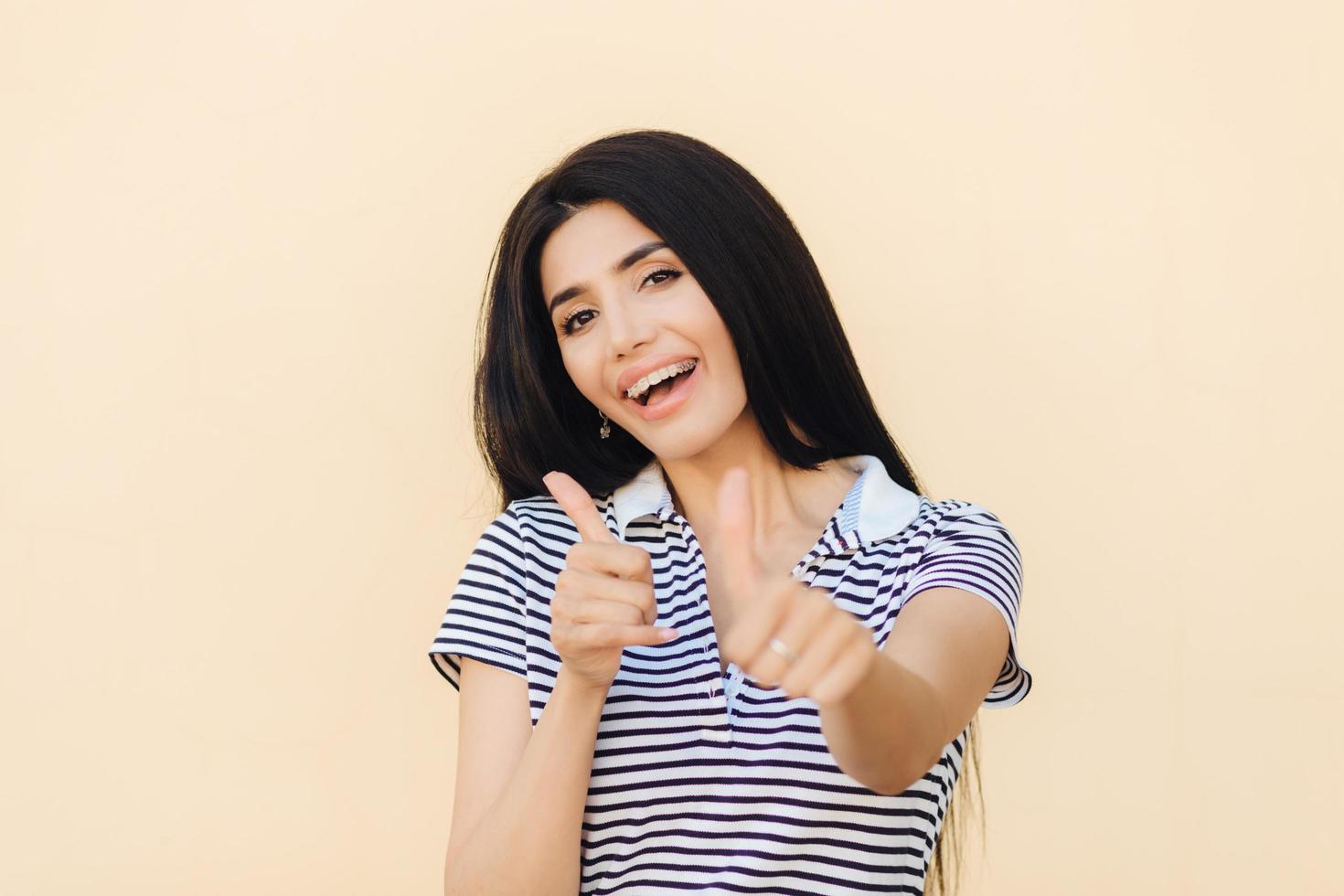 l'inquadratura di una donna europea attraente dà il doppio pollice in su, ha un'espressione positiva, ha i denti bianchi con parentesi, vestita in modo casual, fa un gesto d'approvazione e di approvazione. persone e linguaggio del corpo. foto