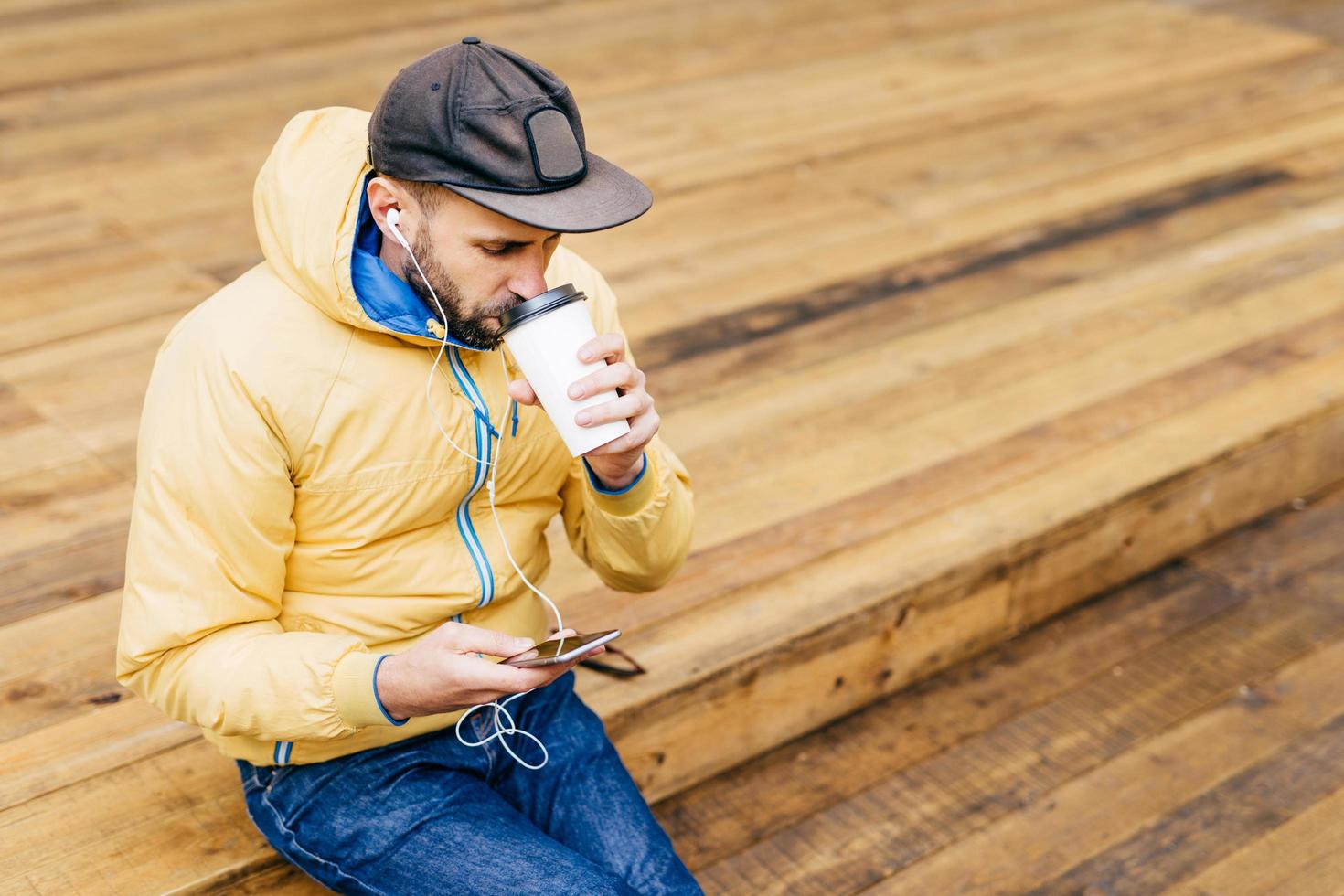 ritratto di uomo alla moda con barba che indossa berretto, giacca gialla e jeans che beve un caffè delizioso e ascolta musica con gli auricolari che tiene il cellulare scaricando foto e video online