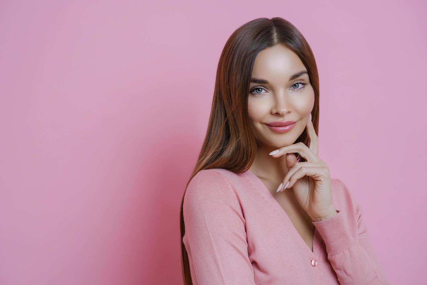 la giovane bella donna europea tocca delicatamente il viso, guarda con espressione compiaciuta alla telecamera, ha una pelle sana e perfetta, ascolta i consigli degli amici, indossa un maglione casual, isolato su sfondo rosa foto