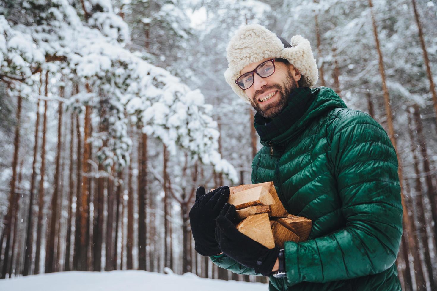 scatto all'aperto di un maschio felice sorridente con barba e baffi che indossa occhiali, giacca a vento e cappello caldo, tiene la legna da ardere, si erge contro alberi coperti di neve, gode di aria fresca e gelida nella foresta foto