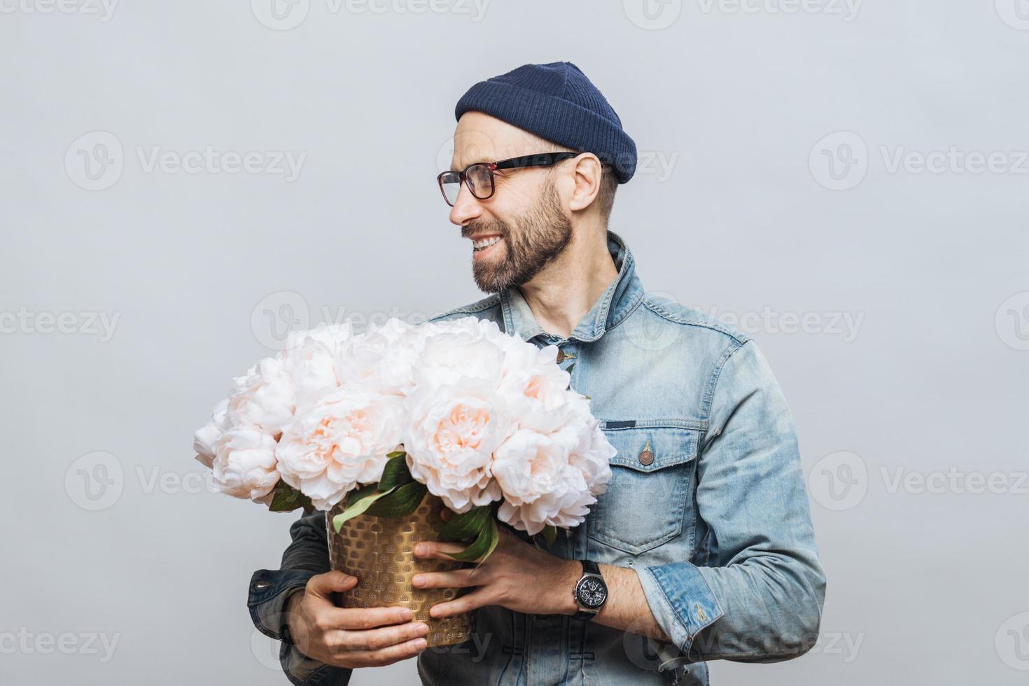 il colpo orizzontale del maschio barbuto felice distoglie lo sguardo mentre tiene un mazzo di fiori bianchi, essendo di buon umore come ha appuntamento con la sua ragazza, le fa un regalo, isolato su sfondo bianco. foto