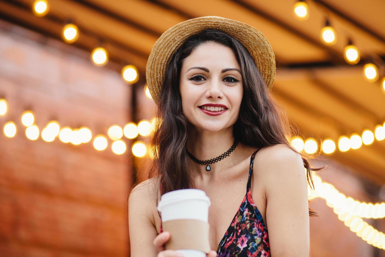 giovane donna bruna in cappello di paglia estivo e vestito, trucco, tenendo una tazza di carta con una bevanda calda, riposando in una caffetteria all'aperto, posando alla telecamera con un sorriso, dimostrando i suoi denti bianchi. foto