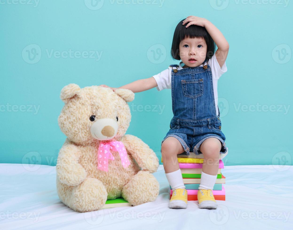 bambino poco asiatico ragazza o un' poco carino asiatico ragazza lettura un' libro nel il verde sfondo e seduta su il libri con un' giocattolo orsacchiotto orso foto