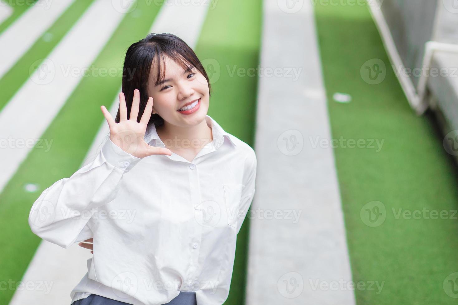 carino giovane asiatico scuola ragazza chi indossa bianca camicia in piedi e sorridente mentre all'aperto nel il città con un erba nel il sfondo. foto